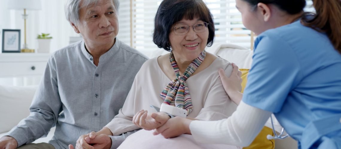 Scrub nurse doctor using digital tonometer check blood pressure for old asia female at home in elderly care cardiovascular medical visit, cholesterol problem and hospice healthcare caregiver concept.